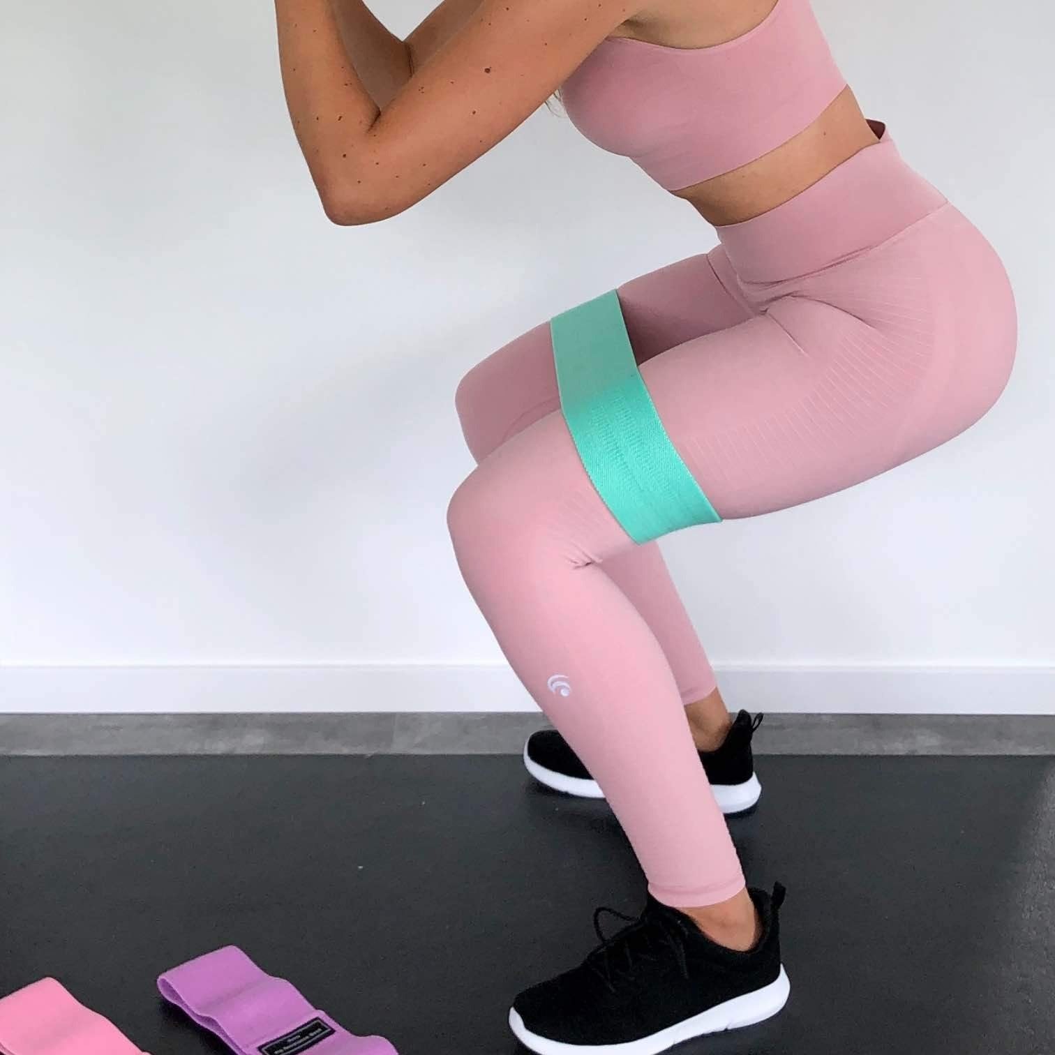Blogger booty bands - accessories Model in all pink gym clothes with black & white sneakers doing a squat using the green Blogger Booty Band around her thighs just above the knees