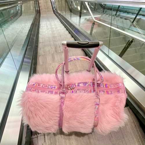 Weekender bag made of pink faux fur and iridescent pink faux leather handles and shoulder strap used as a carry-on bag sitting on a suitcase traveling down an escalator at the airport.