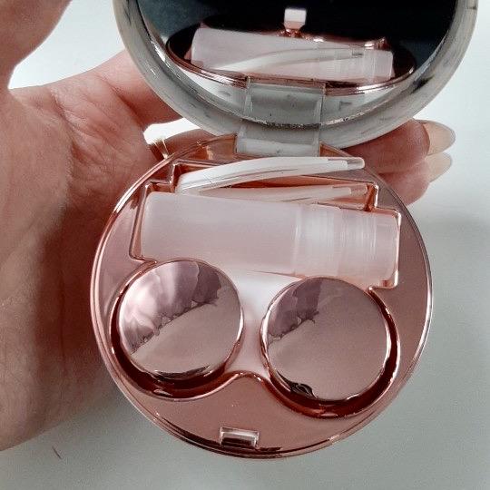 Unity contacts kit - rose gold - accessories. Hand holding open rose gold kit displaying inner lid mirror, solution bottle, lens holder & tweezers in the designated slots for each item.